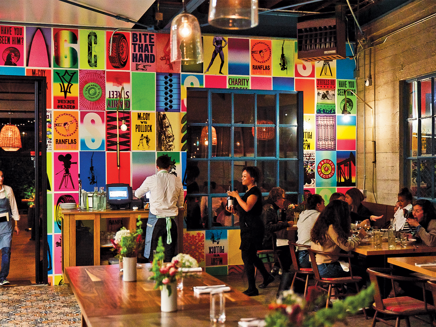 A busy restaurant scene with brightly colored wall paper that has a grid layout with neon tiles with either big letters or graphic images.