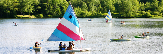 A variety of paddle boats and sail boats with candy striped sails cover the dark green surface of Lake Arax at Camp Nubar.