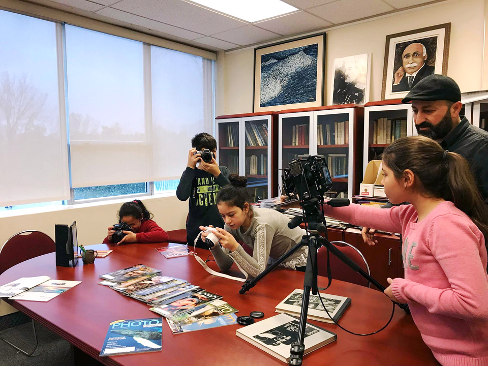Youth in an office setting are learning how to use various types of cameras. Some are filming while others are arranging magazines and other objects for product shots.