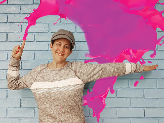 Young girl with a beige sweater and a cap in front of a blue brick wall and a pink splash coming out of her finger