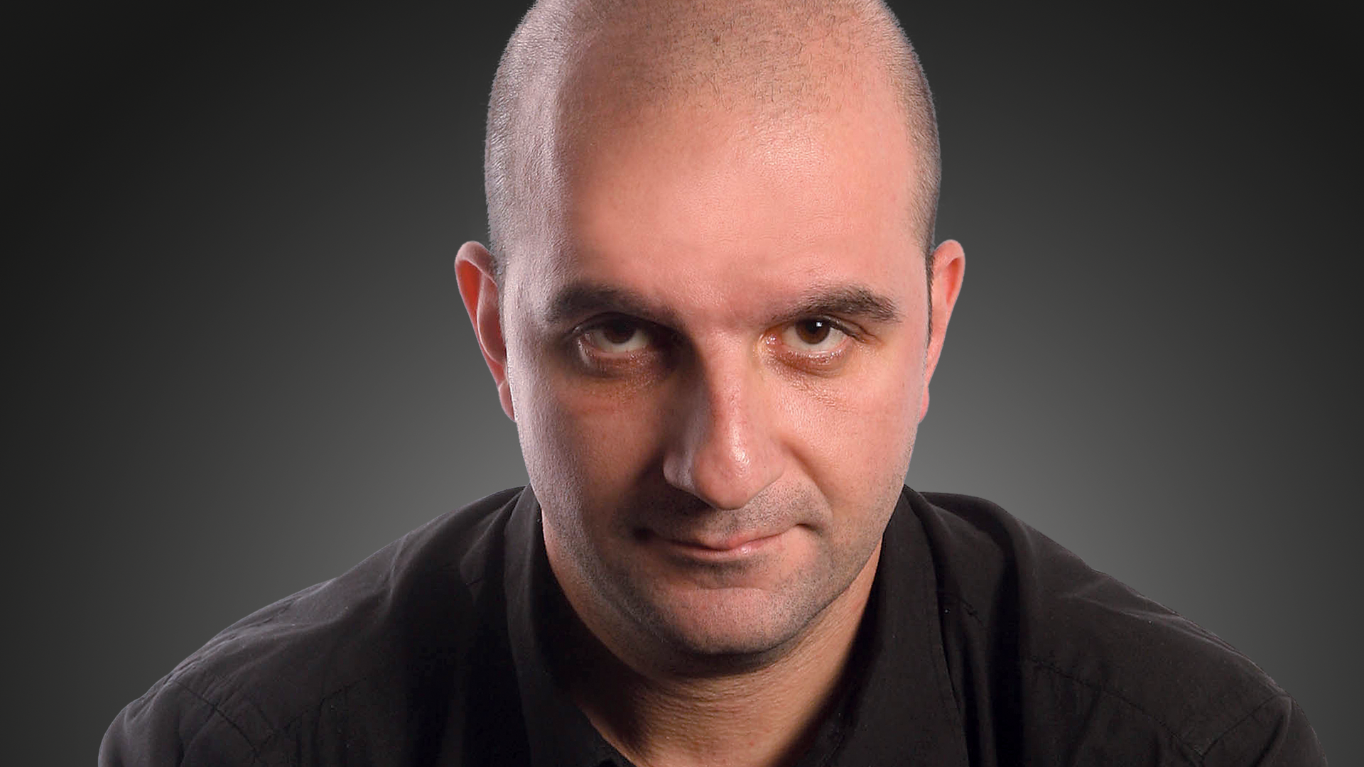 A bald young man looking straight to the camera on a dark background