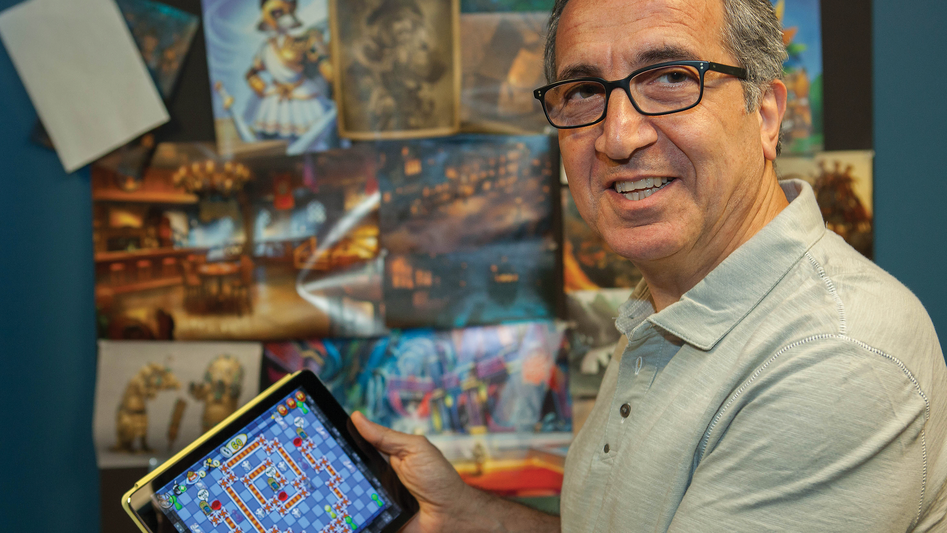 A man with glasses staring at the camera, showing the game he developed on a tablet in his hand.