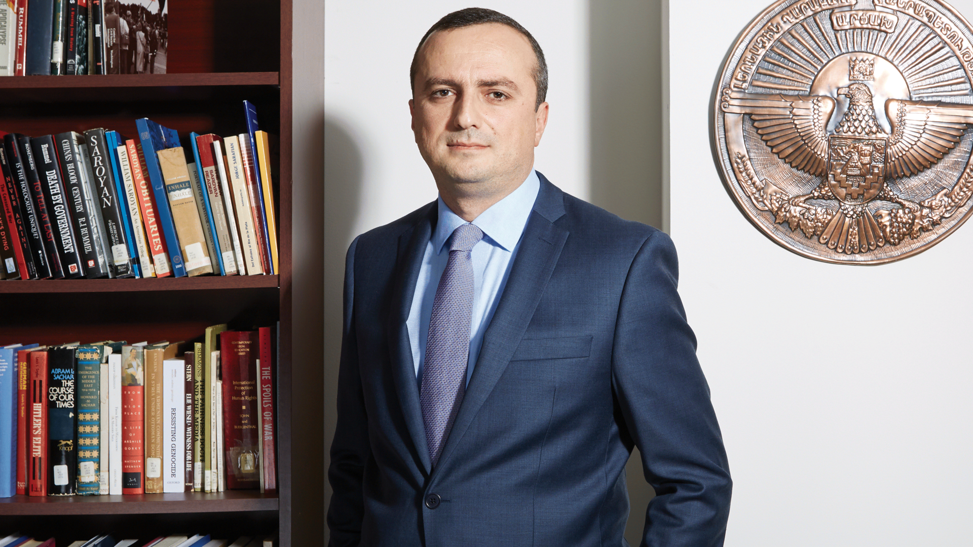 Robert Avetisyan, in a blue suit, stands on the side of a bookshelf.