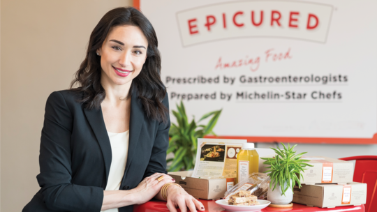 An Armenian woman in a black blazer, smiling and resting her arm on a table with an assortment of gourmet food on it. The woman is smiling and the logo of her company "Epicured" is in the background.