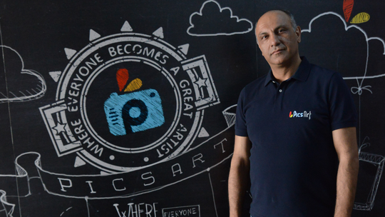 A man staring at the camera, standing in front of a black wall with picsart logo on it.