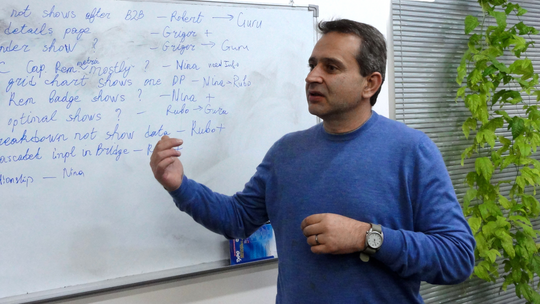 A man with a blue sweater in front of a whiteboard.