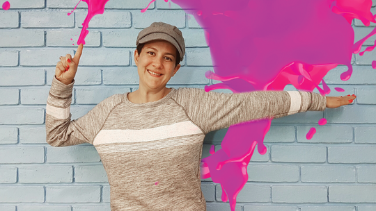 Young girl with a beige sweater and a cap in front of a blue brick wall and a pink splash coming out of her finger
