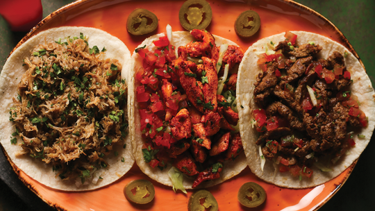Three different types of asado tacos plated on a reflective hand made oblong orange dish.